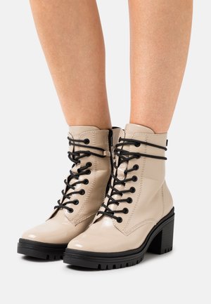 Botas de tobillo de charol beige con cordones negros, un tacón grueso y una suela texturizada. Cuenta con un zipper en el lateral para facilitar su uso.
