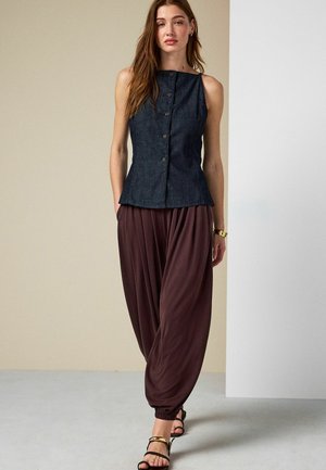 Femme portant un top sans manches foncé avec des boutons, un pantalon marron ample resserré aux chevilles, des sandales noires et un bracelet en or, debout à l'intérieur.