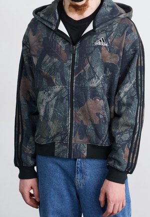 Mann trägt einen Adidas Kapuzenpullover mit Camouflage-Muster, schwarzen drei-Streifen-Ärmeln und blauer Jeans, steht vor einem einfarbigen Hintergrund.