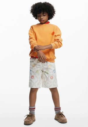 Oranžová mikina s výšivkou kvetov, biele šortky s farebnými vzormi, hnedé topánky a sivé pruhované ponožky. Jednofarebné pozadie.