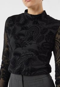 Kobieta ubrana w czarną przezroczystą bluzkę z długim rękawem z aksamitem w paisley wzór i wysokim kołnierzem, zestawioną z szarymi dopasowanymi spodniami.