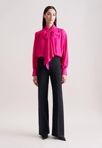 Blusa di raso rosa acceso con un grande fiocco al collo, abbinata a jeans neri a vita alta e a zampa. Tessuto liscio e maniche lunghe.