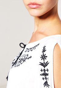 Blouse blanche avec broderies florales noires, un nœud décoratif et des épaules dénudées. Tissu doux avec une coupe décontractée et des détails intriqués.