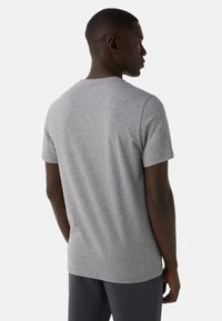 Mężczyzna stojący bokiem, ubrany w szary t-shirt z kr�ótkim rękawem i ciemne spodnie, na neutralnym tle.