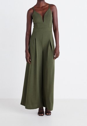 Skokowa kombinezon oliwkowo-zielony z głębokim dekoltem w kształcie litery V, cienkimi ramiączkami, plisami w talii i szeroką nogawką. Gładka tkanina o miękkiej fakturze.