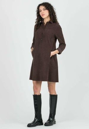 Vrouw met krullend haar, gekleed in een bruine jurk met lange mouwen en knopen, en zwarte kniehoge laarzen, staand met handen in de zakken.