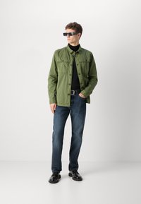 Zelená bavlněná bunda přes černý rolák, spárovaná s modrými džíny a černými botami. Modelka nosí tmavé obdélníkové sluneční brýle.