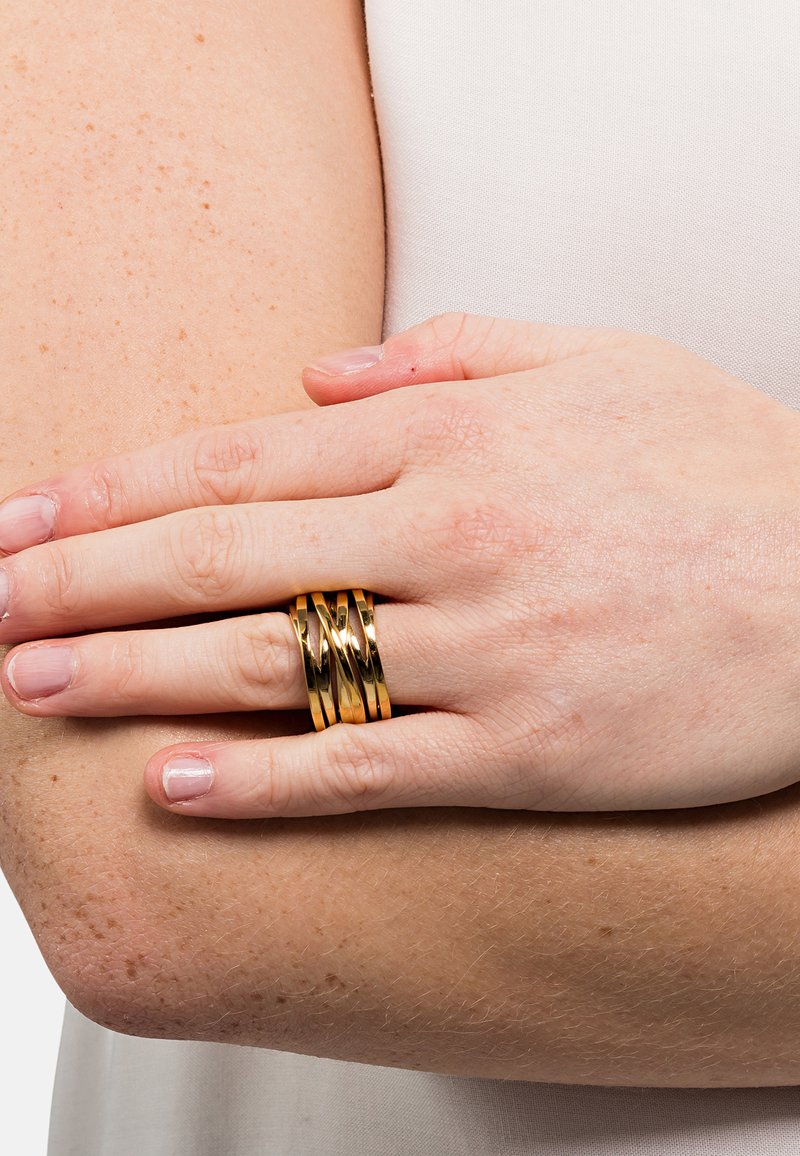 Goldfarbene Ring-Set mit gedrehter und geschichteter Gestaltung. Die Ringe haben eine polierte Oberfläche und werden an einer Hand getragen, die auf einem Arm ruht.