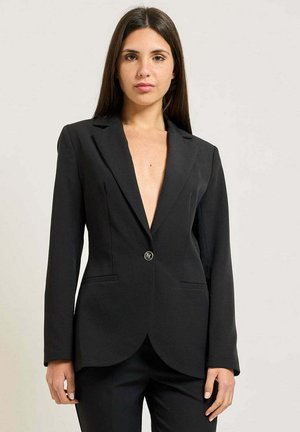 Donna con lunghi capelli scuri indossa un blazer nero aderente con un bottone singolo e pantaloni neri abbinati, in piedi davanti a uno sfondo semplice.