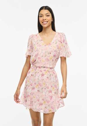 Ženska, ki se smeji v svetlo roza obleki z rožnatim vzorcem, kratkimi zvončastimi rokavi in ožanim pasom, na preprostem belem ozadju.