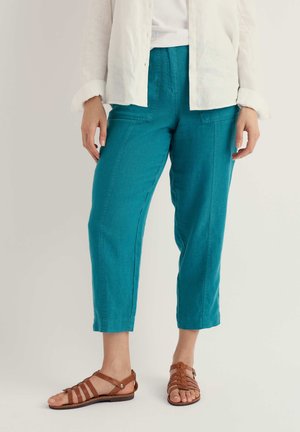 Femme portant un pantalon court couleur sarcelle et des sandales plates marron à brides, avec une chemise blanche à manches longues partiellement visible.