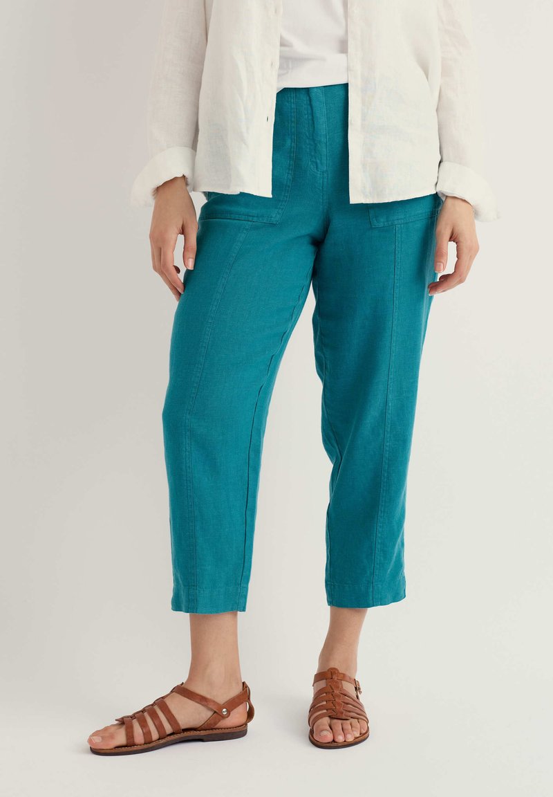 Femme portant un pantalon court couleur sarcelle et des sandales plates marron à brides, avec une chemise blanche à manches longues partiellement visible.