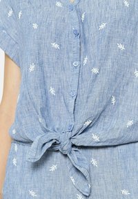 Camisa de botones de color azul claro, hecha de una tela texturizada, que presenta bordados de hojas blancas y un detalle de nudo en el frente. Mangas cortas y diseño de escote en V.