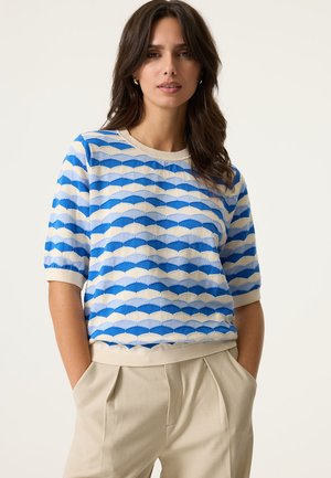 Blue and cream patterned knit sweater with short sleeves and a ribbed neckline, paired with light beige tailored trousers.