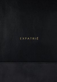 Black leather product featuring a smooth texture with a matte bottom section. Gold embossed "EXPATRIÉ" logo centered on the upper part.