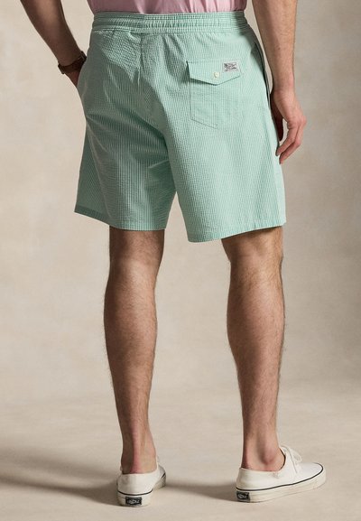 Άνδρας που φοράει πράσινη και λευκή ριγέ βερμούδα με πίσω τσέπη, ροζ πουκάμισο μέσα στη βερμούδα και λευκά παπούτσια slip-on, όρθιος σε μπεζ πάτωμα.