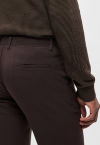 Camisola castanha texturada com trama apertada, combinada com calças castanhas escuras que apresentam um bolso traseiro e tecido suave. O material é macio.