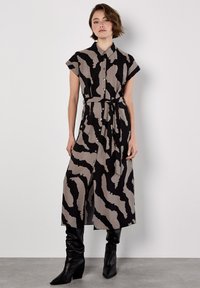 Femme portant une robe midi noire et beige à imprimé abstrait, à boutons, avec des manches courtes, une taille nouée et des bottes à talons noires.