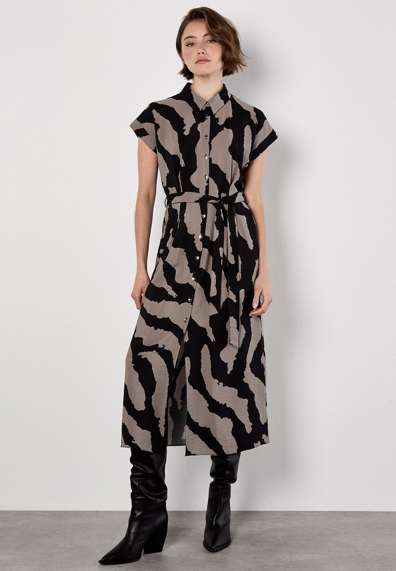Donna che indossa un abito midi a bottoni nero e beige con stampa astratta, maniche corte, vita con nodo e stivali neri con tacco.