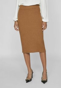 Geribde, bruine midi rok met een aansluitende taille en een zijopening. Draagt met zwarte pumps met een spitse neus en gouden hardware accenten.
