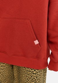 Červená mikina s predným vreckom, rebrovanými manžetami a lemom. Obsahuje malú ružovú nálepku na vrecku. Nosená na leopardích spodkoch.