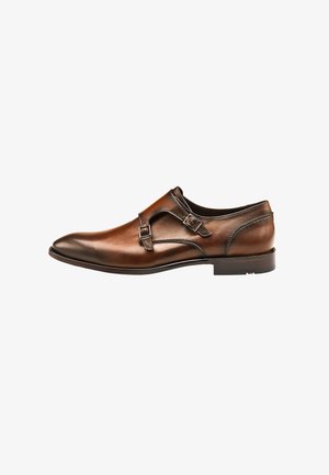 Brown leather shoe with a pointed toe, dual buckles, and a smooth finish. Features a low, stacked heel and subtle stitching details.