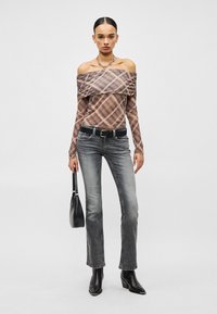 Γυναίκα που φοράει πλεκτό τοπ με ανοιχτούς ώμους και καρό σχέδιο, ξεθωριασμένο γκρι τζιν, μαύρη ζώνη, μαύρα μποτάκια, κρατώντας μαύρη τσάντα, με τα μαλλιά τραβηγμένα πίσω.