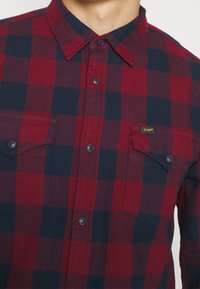 Rød og marineblå ternet flannel skjorte med knap-ned krave, to brystlommer og sorte knapdetaljer. Har et lille "Lee"-logo.