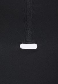 Czarny sportowy top na zamek z teksturowanej tkaniny, z pionowym zamkiem i małą poziomą białą metką w okolicy dekoltu.