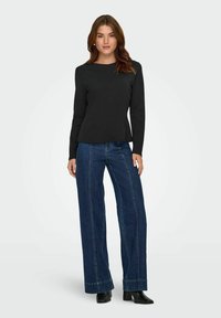 Zwarte langetermijn top met een ronde hals, gecombineerd met wijde blauwe jeans met contrasterende naden. Model staat zelfverzekerd.