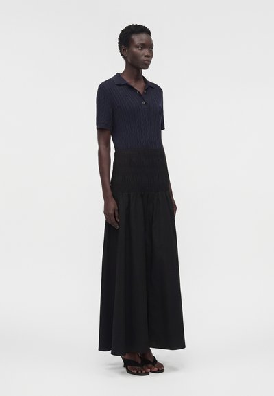 Γυναίκα φοράει μπλε σκούρο πλεκτό τοπ με κοντά μανίκια και γιακά, συνδυασμένο με μαύρη μακριά φούστα ψηλόμεση και μαύρα πέδιλα με ανοιχτά δάχτυλα.