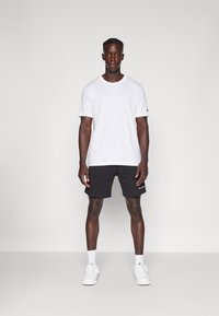 White cotton T-shirt with a crew neckline paired with black shorts. The shorts feature embroidered branding. Footwear is white sneakers.