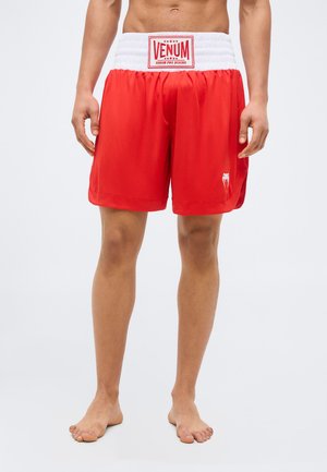 Άνδρας που φοράει κόκκινα μποξεράκια Venum με λευκή μέση, ξυπόλητος, στέκεται σε απλό φόντο.