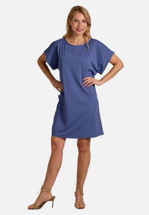 Woman standing with hands on hips wearing a short-sleeve, knee-length blue dress and beige high-heeled sandals against white background.