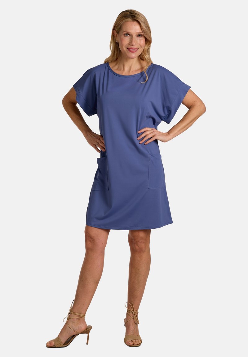 Woman standing with hands on hips wearing a short-sleeve, knee-length blue dress and beige high-heeled sandals against white background.