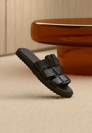 Sort læder slid-sandal med brede remme og riller i sålen på beige tæppe nær brunt rundt sofabord.