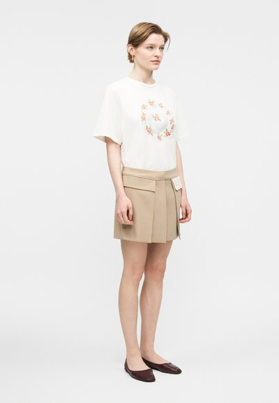 Ung kvinne står i beige plissert skjørt, hvit blomstrete t-skjorte og brune flate sko mot en ensfarget hvit bakgrunn.