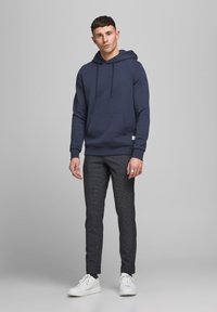 Hoodie bleu marine avec poche avant, poignets côtelés assortis, associé à un pantalon texturé sombre et des baskets blanches. Design simple et décontracté.