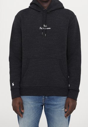 Mand iført en mørkegrå hættetrøje med hvid "Polo Ralph Lauren" skrift på brystet, hvidt logo på ærmet, for- lomme, kombineret med blå jeans.