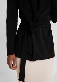 Czarna kurtka zapinana na wiązanie w talii, z długimi rękawami, wykonana z miękkiego materiału, w połączeniu z beżową spódnicą w prążki.