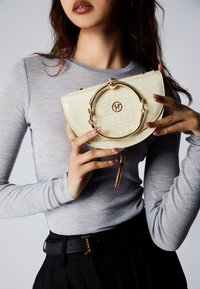 Borsa a manico circolare dorato e dettagli con nappina, in pelle sintetica color crema con motivo coccodrillo, tenuta da una persona con una maglietta grigia a maniche lunghe.