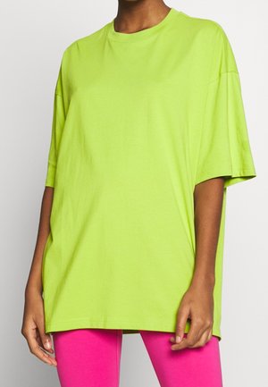 Henkilö, joka pukeutunut ylisuureen kirkkaan limenvihreään T-paitaan, jossa on lasketut olkapäät, ja kuumiin pinkkeihin leggingsseihin yksinkertaista taustaa vasten.