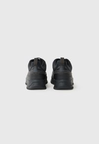 Une paire de baskets de sport noires avec des languettes au niveau du talon, vues de derrière sur un fond blanc uni.