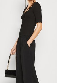 Haut noir côtelé à manches courtes avec une coupe ajustée, bords festonnés et fermeture à boutons, assorti à un pantalon noir taille haute et un petit sac à main.