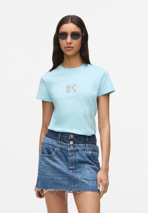 Žena v okrúhlych čiernych slnečných okuliaroch, svetlomodrom tričku s malým logom a vysoko pásovej denimovej mini sukni s gombíkovými detailmi.