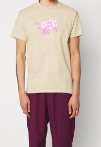 Beige bomulls t-shirt med korta ärmar. Har en rosa grafik av ett bi och ordet "OBEY" ovanför, med stjärnaccent. Normal passform.