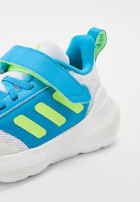 Baskets de sport bleues avec un dessus en maille, présentant un accent vert vif et une sangle, une semelle blanche et des rayures latérales texturées.