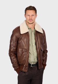 Hnědá kožená bombarďák s krémovým beránkovým límcem, předními kapsami, vroubkovanými manžetami a zapínáním na zip. Nosený přes olivové tričko.