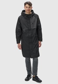 Svart vattentålig parka med huva, med en ficka på bröstet med dragkedja, sidofickor och justerbara snören. Kompletterad med grå jeans och svarta skor.