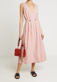 Rosa linneklänning med djup V-ringning, plisserad kjol och två framknappar. Stylad med en röd handväska och svarta slidesandaler med nitar.